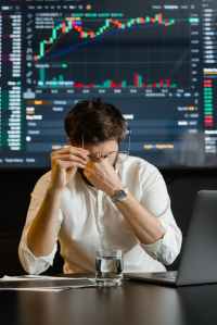 A stressed man in a white shirt sits at a desk with a laptop, holding his glasses and resting his head on his hand. In the background, a financial graph displays market trends.